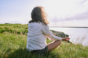Rear view of curly-haired woman sitting with crossed legs on grass and meditating in silence by tranquil lake