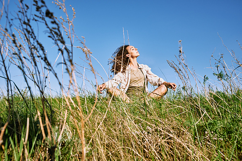 Young relaxed woman with dark long wavy hair crossing legs while sitting in green grass against blue sky and enjoying rest on sunny day