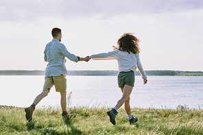 Rear view of affectionate young couple of hikers holding hands and running to lake while enjoying vacation