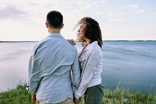 Young serene female with dark curly hair standing close to her husband while both having rest in front of large lake against cloudy sky
