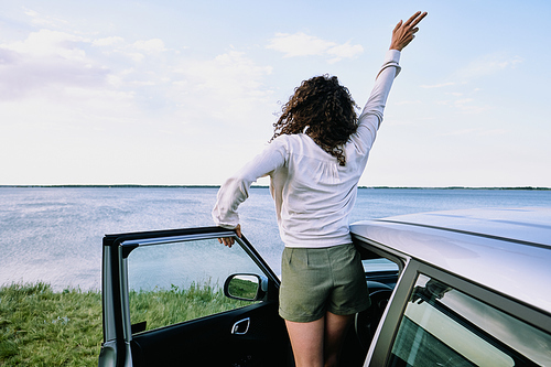 Back view of joyful young woman in casualwear coming out of car and raising her hand while standing in front of lake or river