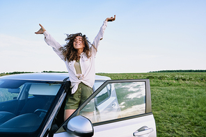 Ecstatic curly-haired girl in casual shirt raising arms and standing on car sill while having fun during car trip