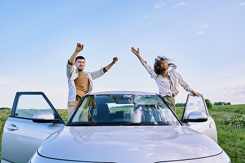 Cheerful excited young couple standing on car sills and waving arms while greeting nature with cries of joy