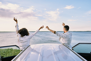 Rear view of young cheerful couple in shirts standing close to their car with open doors and expressing joy of freedom with raised hands
