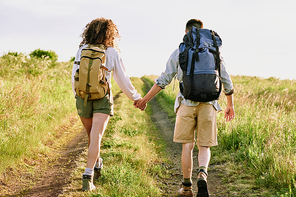 Rear view of young backpackers in shorts holding hands and walking over country road