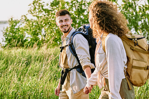 Positive young couple with backpacks holding hands and looking at each other while enjoying hike in country