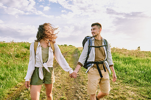 Young amorous couple in casualwear talking while moving down country road against cloudy sky on summer day and looking at one another