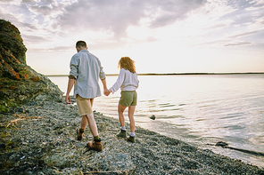 Young amorous hikers in casualwear holding by hands while moving along coastline by water with cloudy sky above and enjoying romantic time