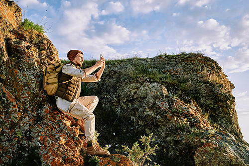 Handsome bearded hiker in vest sitting on cliff rock and photographing landscape on smartphone
