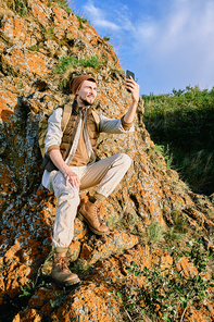 Contemporary young backpacker with rucksack looking in smartphone camera while making selfie on stone rock in the country