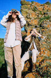 Young woman using binoculars while her boyfriend photographing landscape on smartphone, they enjoying beauty of nature