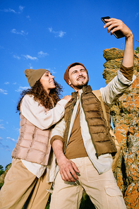 Happy young man with smartphone making selfie with his wife standing near by on background of blue sky and stone rock