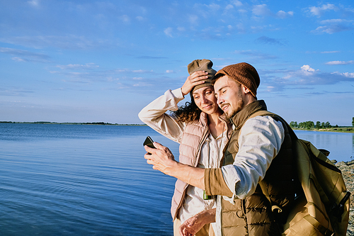 Content beautiful young couple in hats and vests standing at lake shore and taking selfie on smartphone during hike