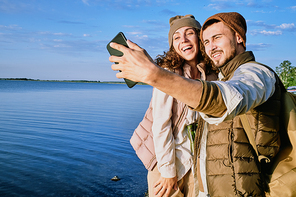 Young cheerful dates standing by each other and looking at smartphone camera while making selfie on background of blue sky and water