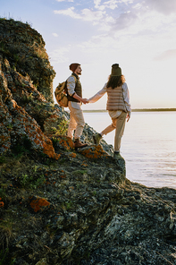 Affectionate hikers in casual clothes standing on rocks of cliff and looking at lake while walking together at sunset