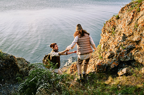 Young bearded man in hat giving hand to girlfriend while assisting her to go down cliff
