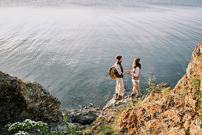 Young affectionate romantic couple holding by hands while standing on stone rock by waterside and looking at one another