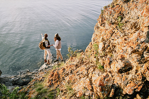 Above view of young man with backpack holding hand of girlfriend and showing lake to her during hiking travel