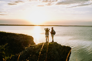 Young male tourist with backpack explaining something to his wife or girlfriend while both standing on top of rock by water at sunset