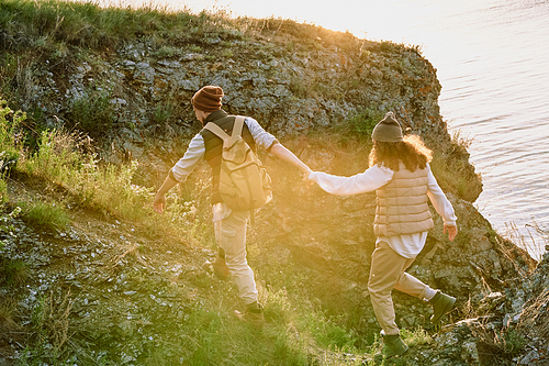 Young male hiker with backpack holding his wife by hand while both moving down stone rock covered by green grass at sunset