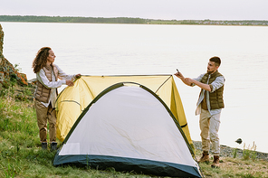 Contemporary young hikers putting tent on the ground by waterside and stretching textile roof over top while preparing for night