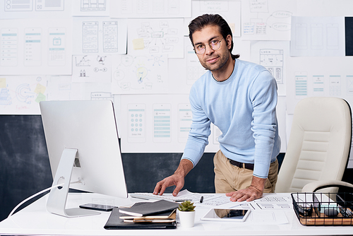 Portrait of confident middle-aged man with stubble standing at desk in office and using computer to check ui design