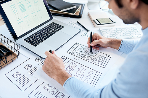 Close-up of busy designer sitting at table with laptop and drawing icons placement for ui design
