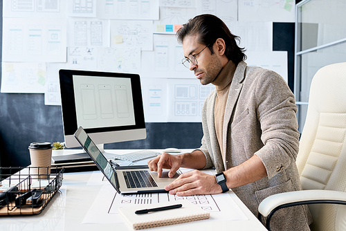 Horizontal shot of handsome man wearing eyeglasses sitting at office desk working on laptop