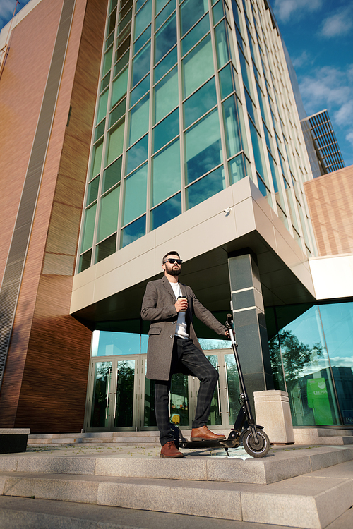 Young elegant businessman having coffee while keeping one leg on electric scooter against large contemporary office building