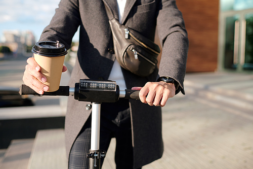 Hands of elegant businessman in coat holding glass of coffee and riding on electric scooter in urban environment while hurrying to work
