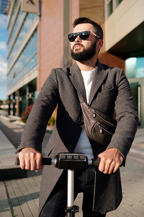 Young elegant businessman in coat and sunglasses riding on electric scooter in urban environment against modern office building