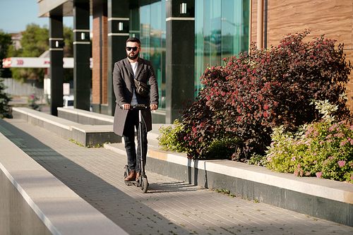 Elegant businessman in sunglasses moving on electric scooter along bushes growing by modern office center while going to work