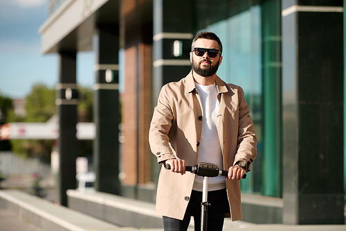 Serious bearded businessman in beige trenchcoat and sunglasses holding by handles of electric scooter while standing against office center