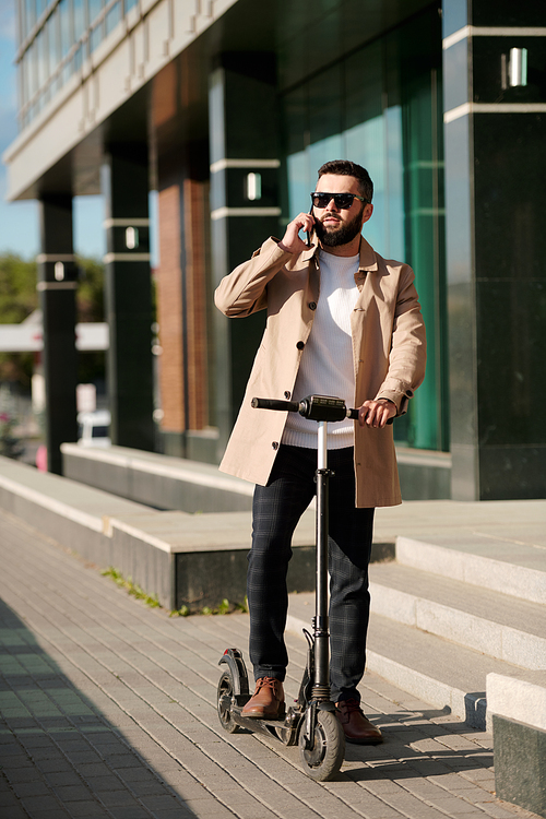 Busy young man in smart casualwear and sunglasses talking on mobile phone outdoors while standing on electric scooter by building