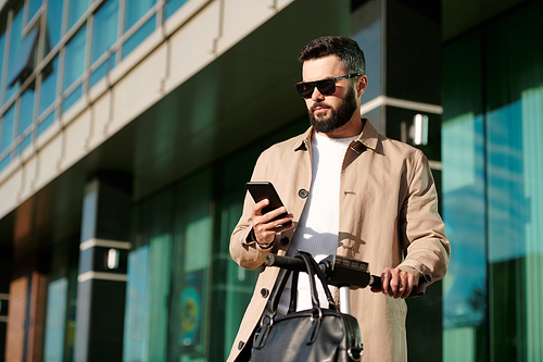 Serious young businessman in beige trenchcoat and sunglasses holding by handles of electric scooter while scrolling in smartphone outdoors