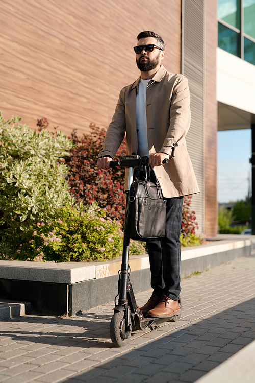 Serious young businessman in elegant casualwear and sunglasses moving on electric scooter along wall of contemporary business center
