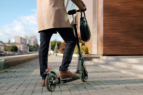 Low section of elegant businessman in beige trenchcoat and pants holding by handles of electric scooter while standing against office building