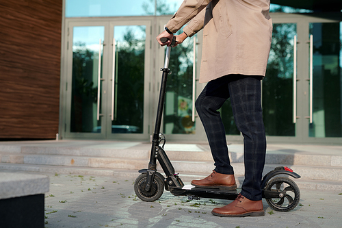 Low section of businessman in trenchcoat and pants holding by handles of electric scooter while standing against entrance of office center