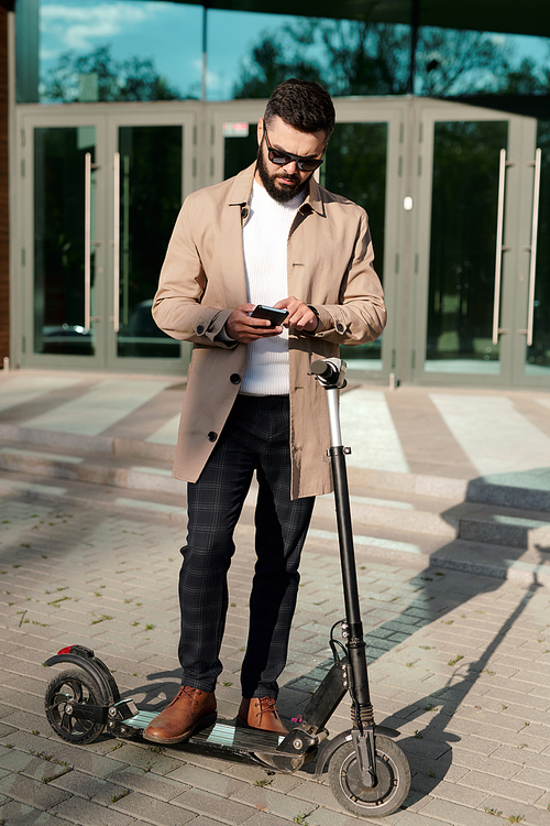 Contemporary young elegant businessman scrolling or texting in smartphone while standing on electric scooter outdoors
