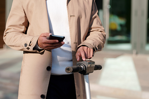 Mid-section of businessman in beige trenchcoat and white knitted pullover scrolling in smartphone while touching panel on electric scooter
