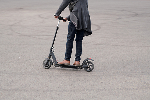 Low section of young contemporary businessman in casualwear standing on electric scooter while moving down asphalt road to work