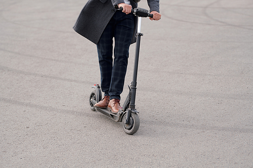 Low section of contemporary businessman in coat and pants riding electric scooter along asphalt road while moving to work in the morning