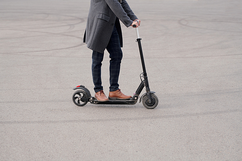 Low section of young contemporary businessman in coat, pants and boots standing on electric scooter while moving to work in the morning