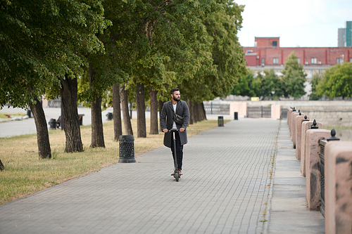 Contemporary young businessman in smart casualwear riding electric scooter along riverside and park in urban environment