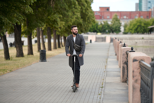 Contemporary young bearded businessman in coat riding on electric scooter in urban environment along riverside in the morning