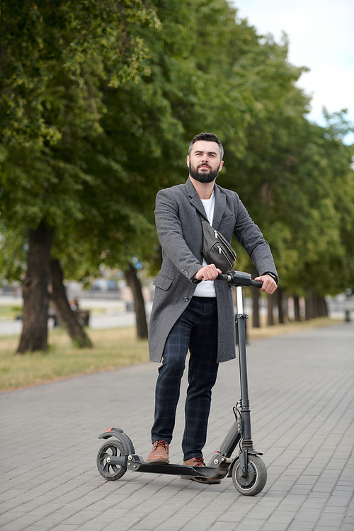 Contemporary young elegant businessman standing on electric scooter in urban environment against row of green trees growing along road
