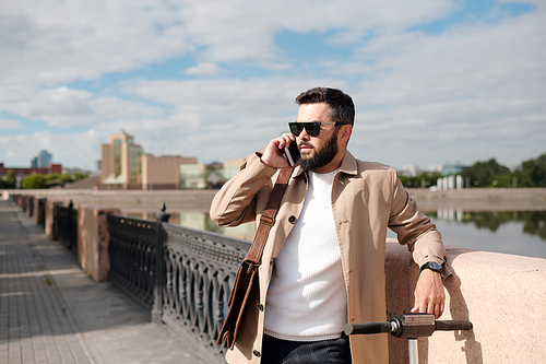 Bearded businessman in beige trenchcoat, white knitted pullover and sunglasses talking on smartphone while standing by riverside