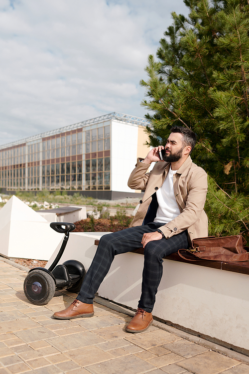 Young bearded busy man in elegant casualwear sitting by coniferous tree in urban environment and talking on smartphone against building