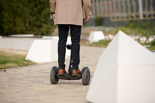Back view of young contemporary man in pants and trenchcoat standing on gyroscope and moving to business center in the morning