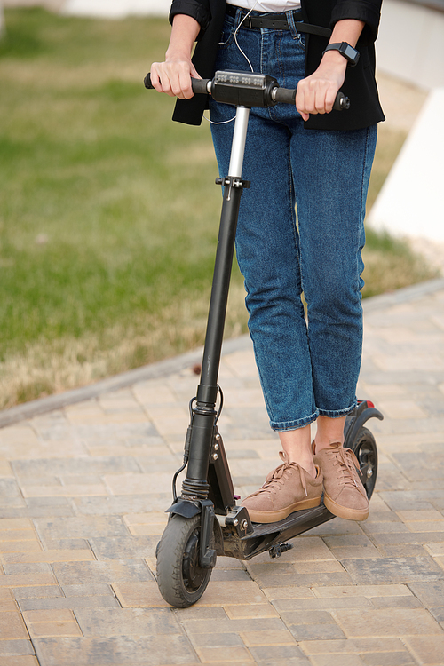 Contemporary young businesswoman in casualwear standing on electric scooter in urban environment against road and green lawn